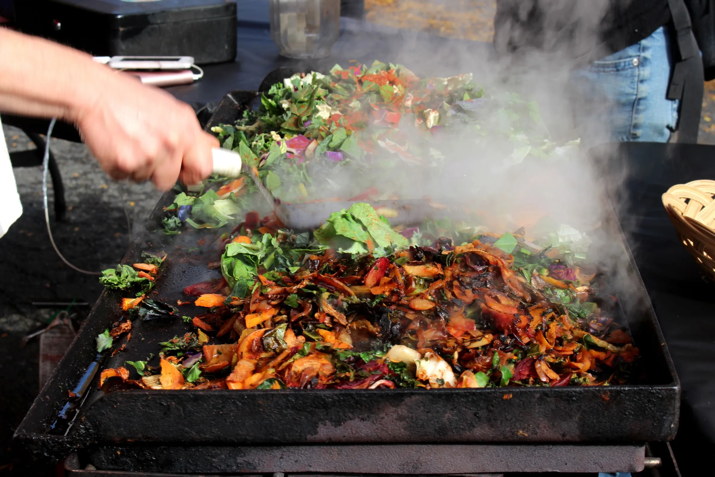 A market griddle piled with chard, carrots and peppers, steam rising as a cook adds a fresh handful of greens.